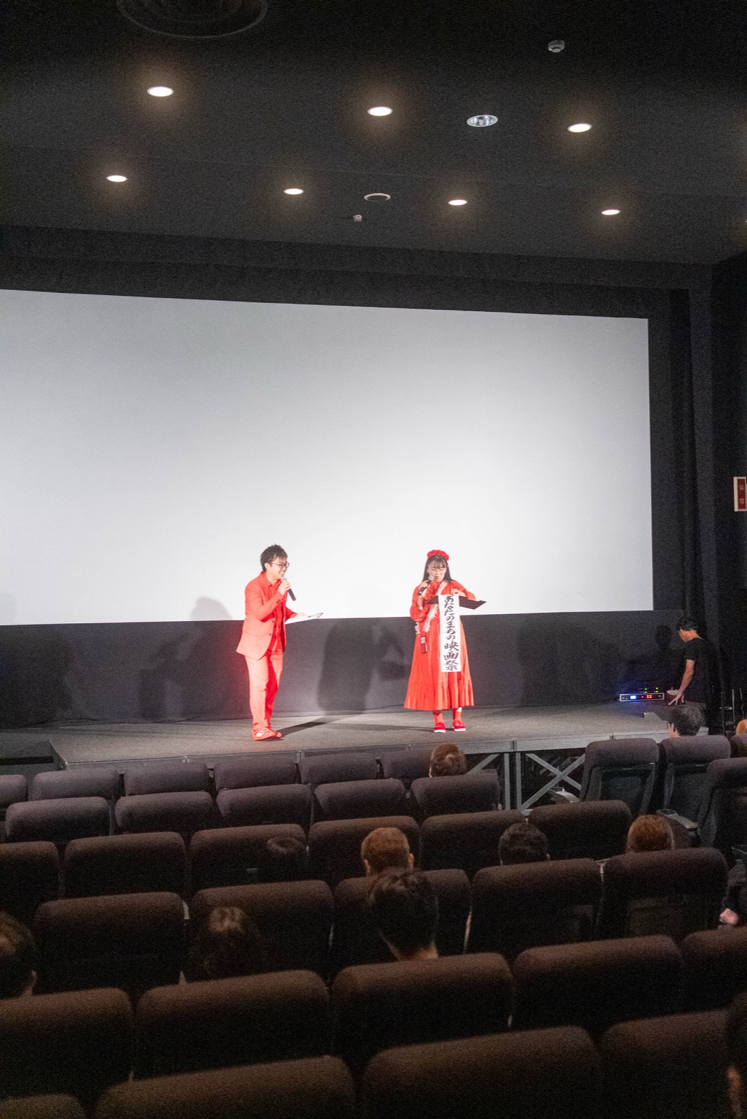 開演前、スクリーン前のステージであいさつする司会